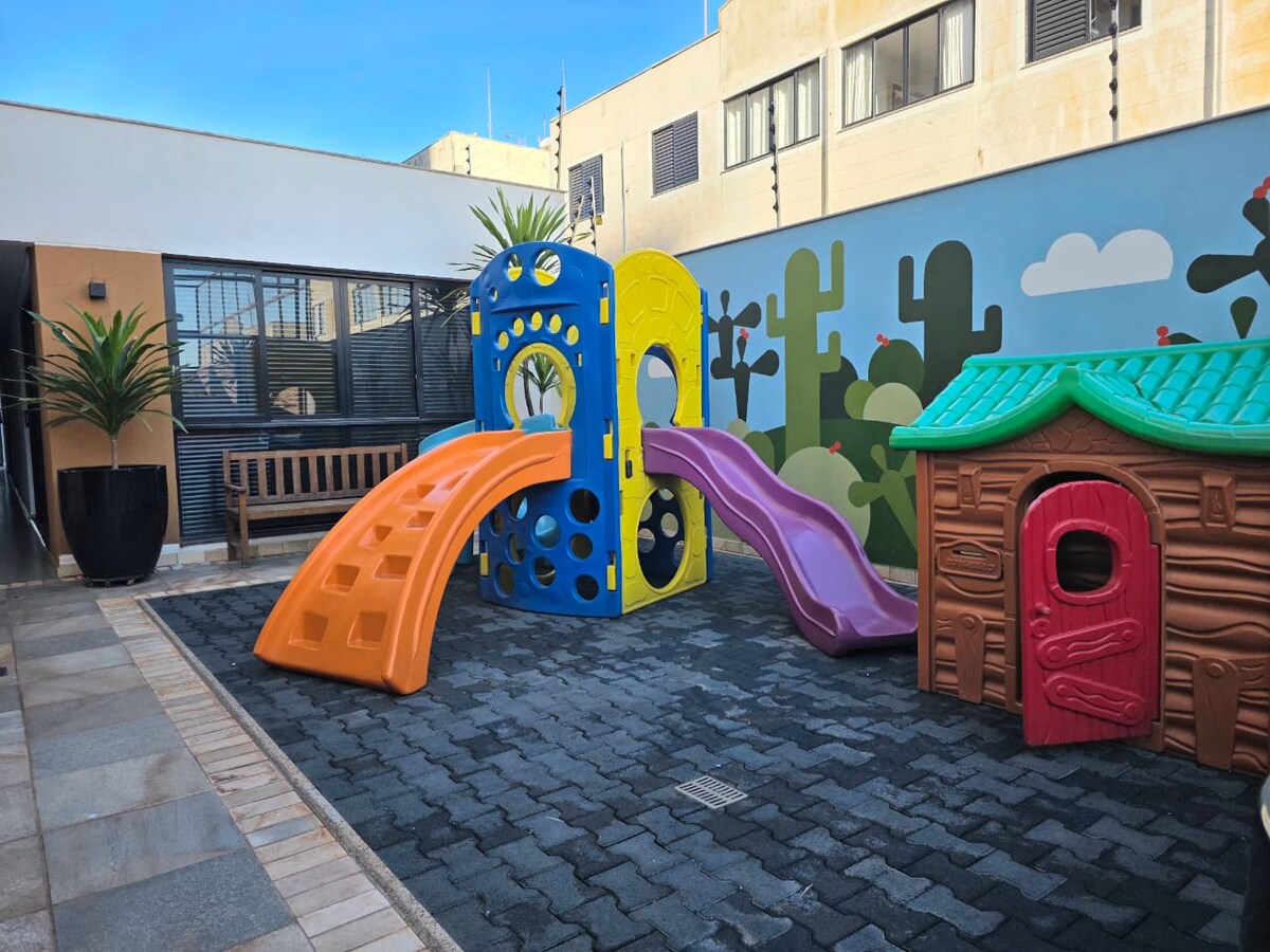 A playful outdoor area features a colorful playground set with a slide, climbing structure, and a small playhouse. Bright green plants are positioned nearby, and a wooden bench provides seating. A mural of cacti adds a playful backdrop to the space.