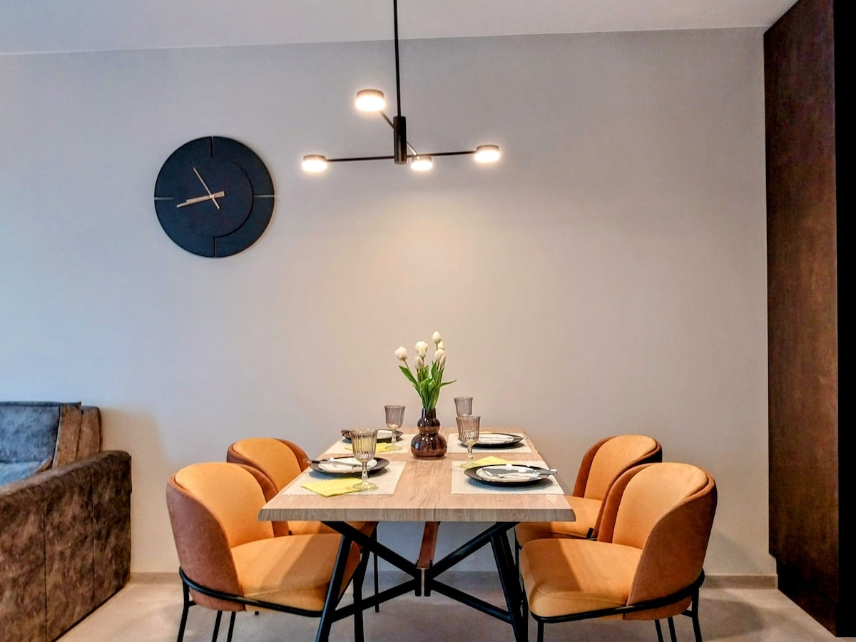 A dining area is featured with four soft chairs surrounding a rectangular wooden table, set with plates and glasses. A simple vase holds fresh flowers at the center. A modern clock adorns the wall above, complemented by soft, neutral tones throughout the space.