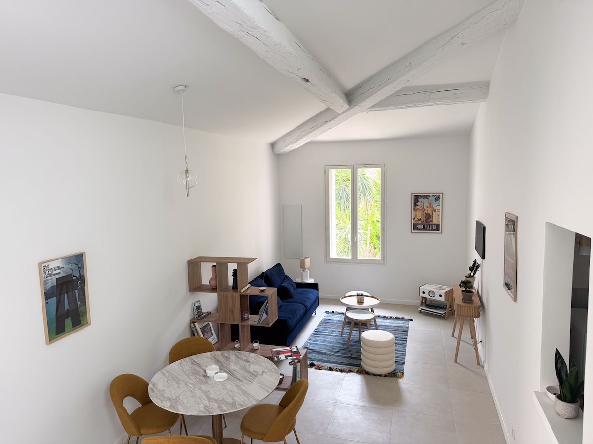A spacious living area is illuminated by natural light from large windows. Wooden beams adorn the ceiling, adding a touch of character. A blue sofa is paired with a round marble table and four mustard-colored chairs, inviting relaxation. A cozy rug enhances the room's inviting atmosphere.