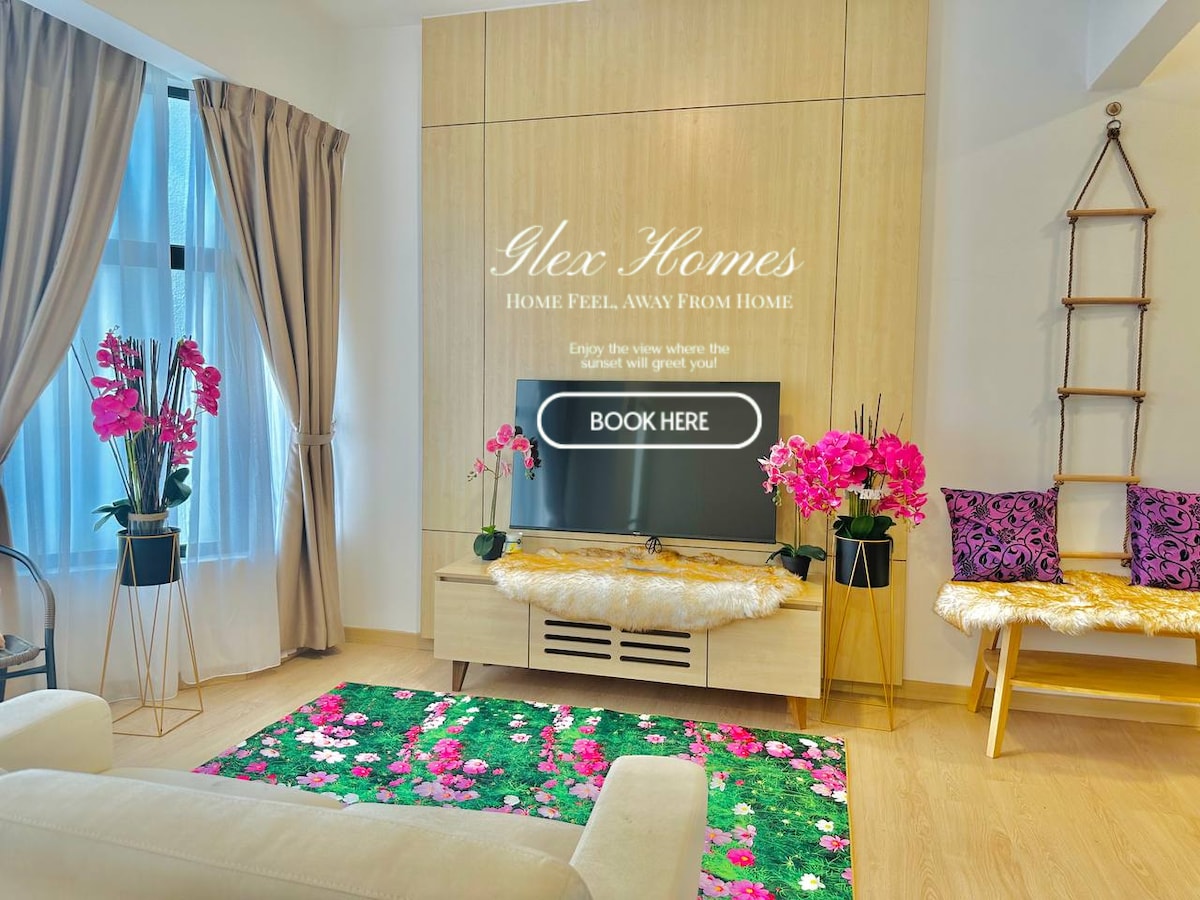 A bright living area is showcased, featuring a low wooden entertainment unit with a television placed above. A green rug adorned with colorful floral patterns is centered on the floor, complemented by elegant orchids in planters. Large windows are framed by soft curtains, allowing natural light.