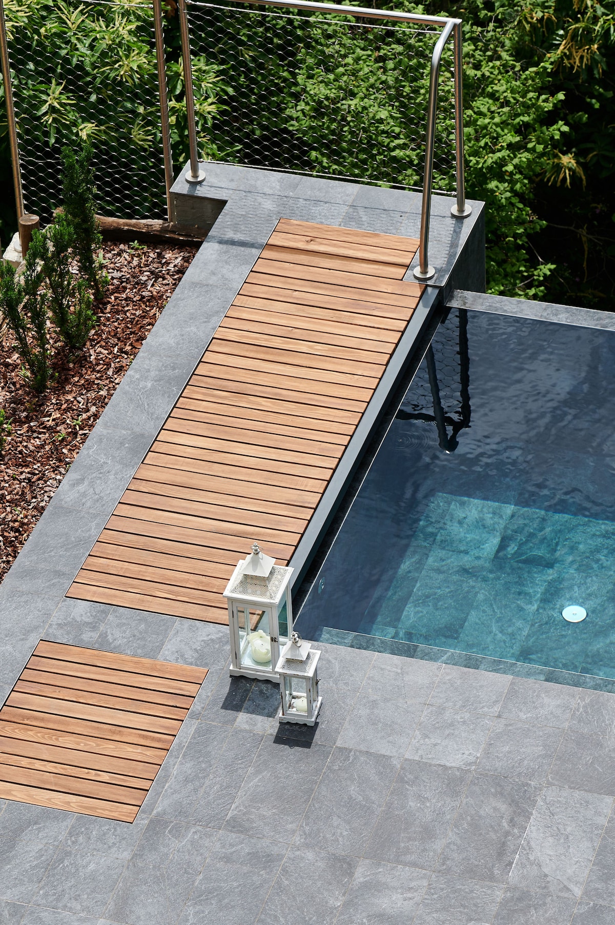 A wooden walkway leads to the edge of a swimming pool, bordered by smooth grey tiles. Two white lanterns are placed on the side, complementing the natural greenery in the background, creating a serene atmosphere around the pool area.