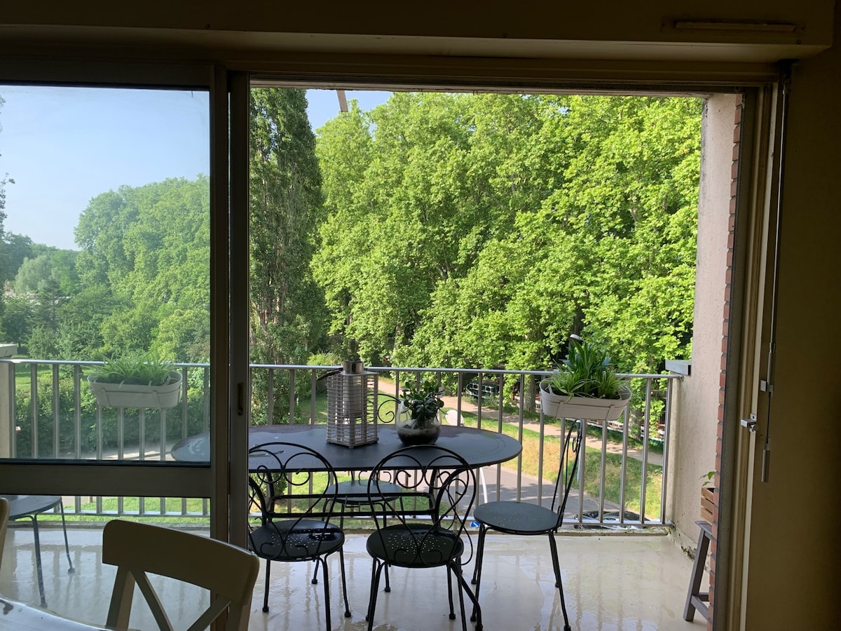 A terrace area is presented, featuring a round dining table surrounded by several chairs. Lush green trees are visible in the background, providing a serene view. Light filters in through the expansive sliding glass doors, enhancing the connection between indoors and outdoors.