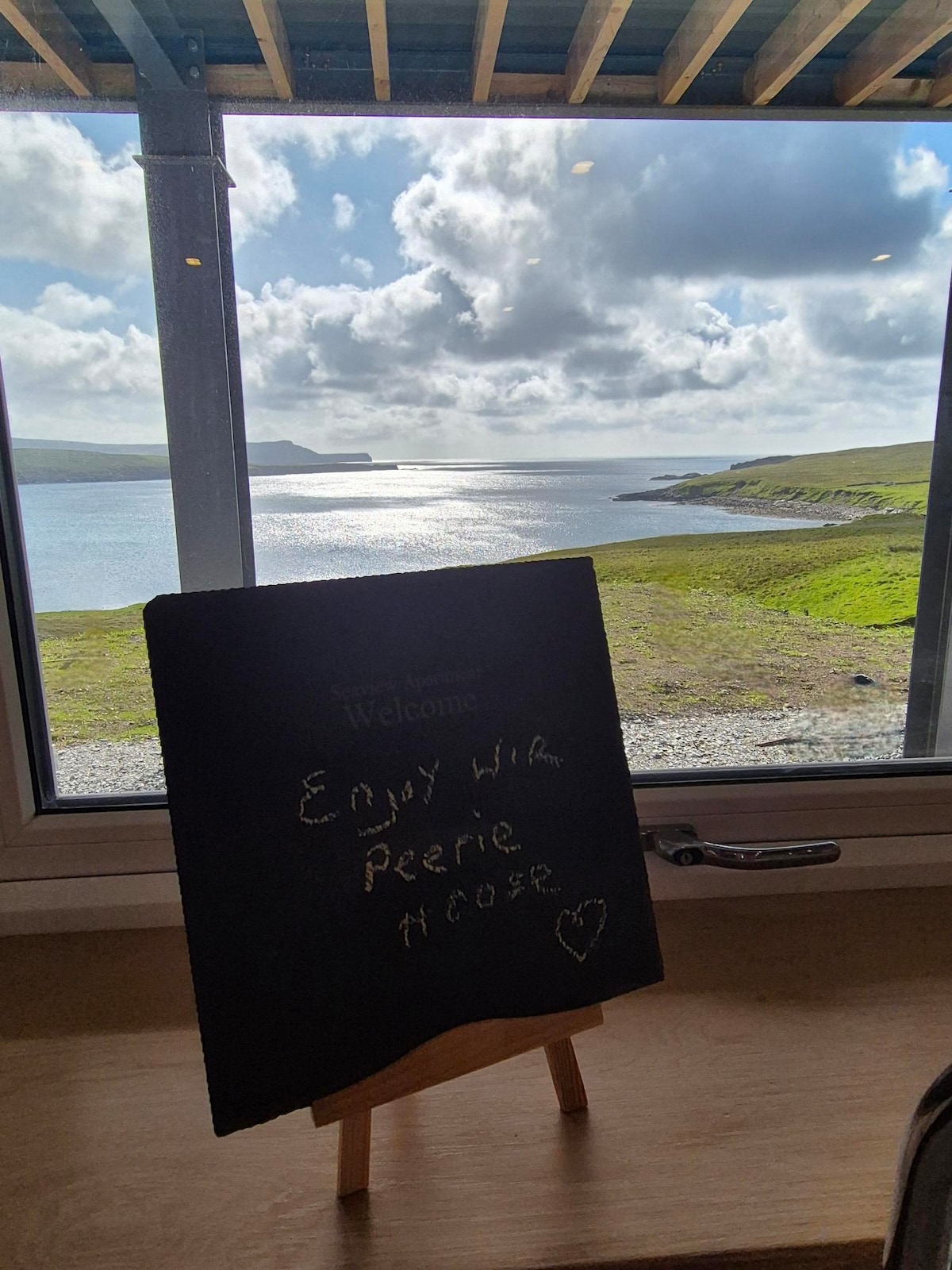 A welcoming chalkboard message stands in front of a large window, framing a scenic view of the water and coastline. The serene landscape features gentle hills under a partly cloudy sky, inviting guests to enjoy the surrounding beauty.