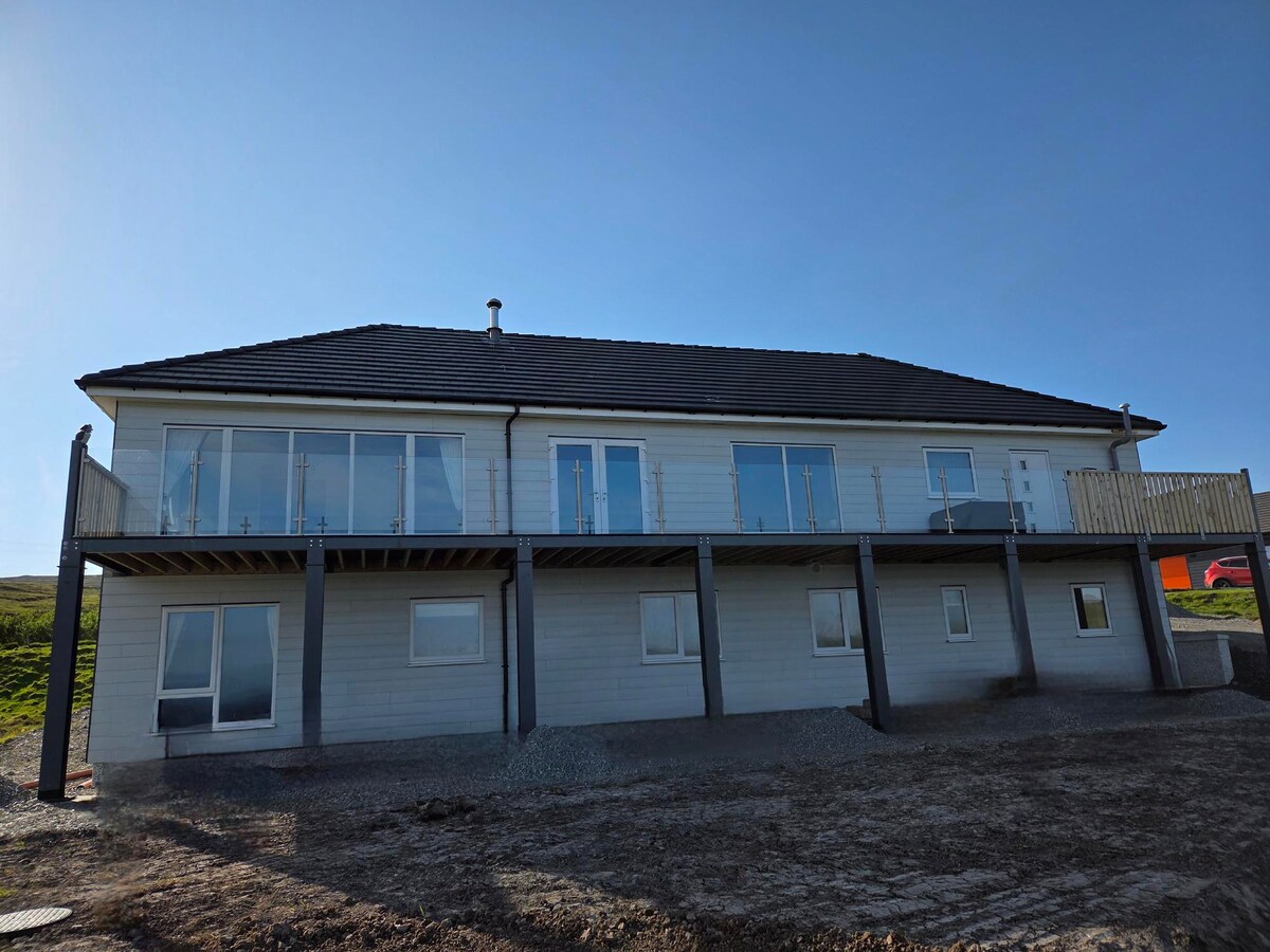 The exterior of the modern apartment is depicted, featuring a two-story structure with large windows that provide natural light. A deck extends from the second floor, offering additional outdoor space. The surrounding area consists of gravel, and the landscape is open and expansive.