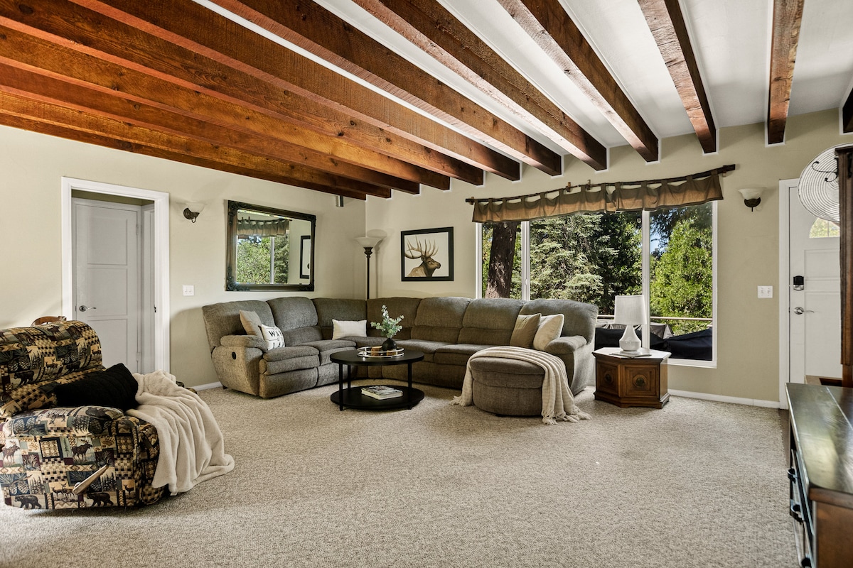 A spacious living room features exposed wooden beams and ample natural light from large windows. Comfortable seating is arranged around a central coffee table, with a plush throw blanket draped over a chair. A cozy ambiance is provided for relaxation.