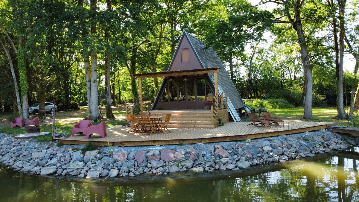 A charming cabin is nestled among tall trees beside a tranquil body of water. The wooden terrace features dining furniture and lounge chairs, providing outdoor seating options. Natural rocks outline the shoreline, creating a peaceful ambiance perfect for relaxation.