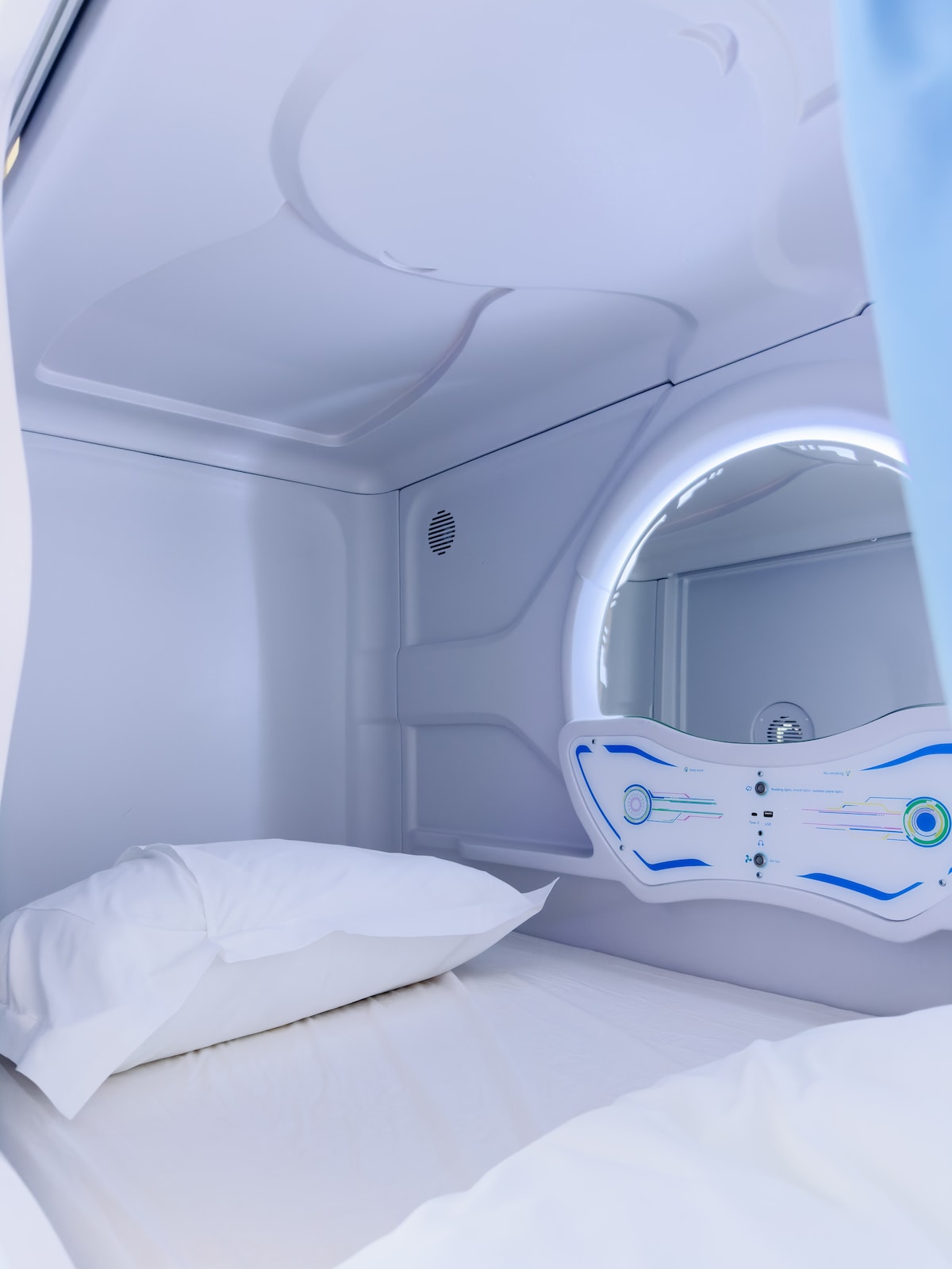 A modern sleeping pod features smooth, curved walls in a subtle white finish. A circular mirror is illuminated by soft light, and a pair of neatly arranged white pillows rest on a light-colored bedding. The space is compact and designed for comfort.