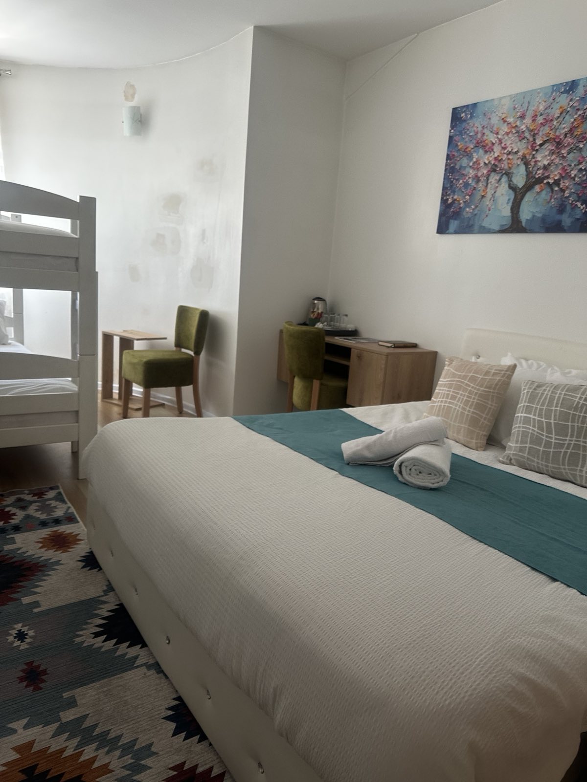 A well-appointed room features a spacious bed covered with crisp linens and a turquoise throw. In the background, a bunk bed is visible. A colorful area rug adds texture, while a small desk and two chairs create a functional space. A piece of artwork adorns the wall.