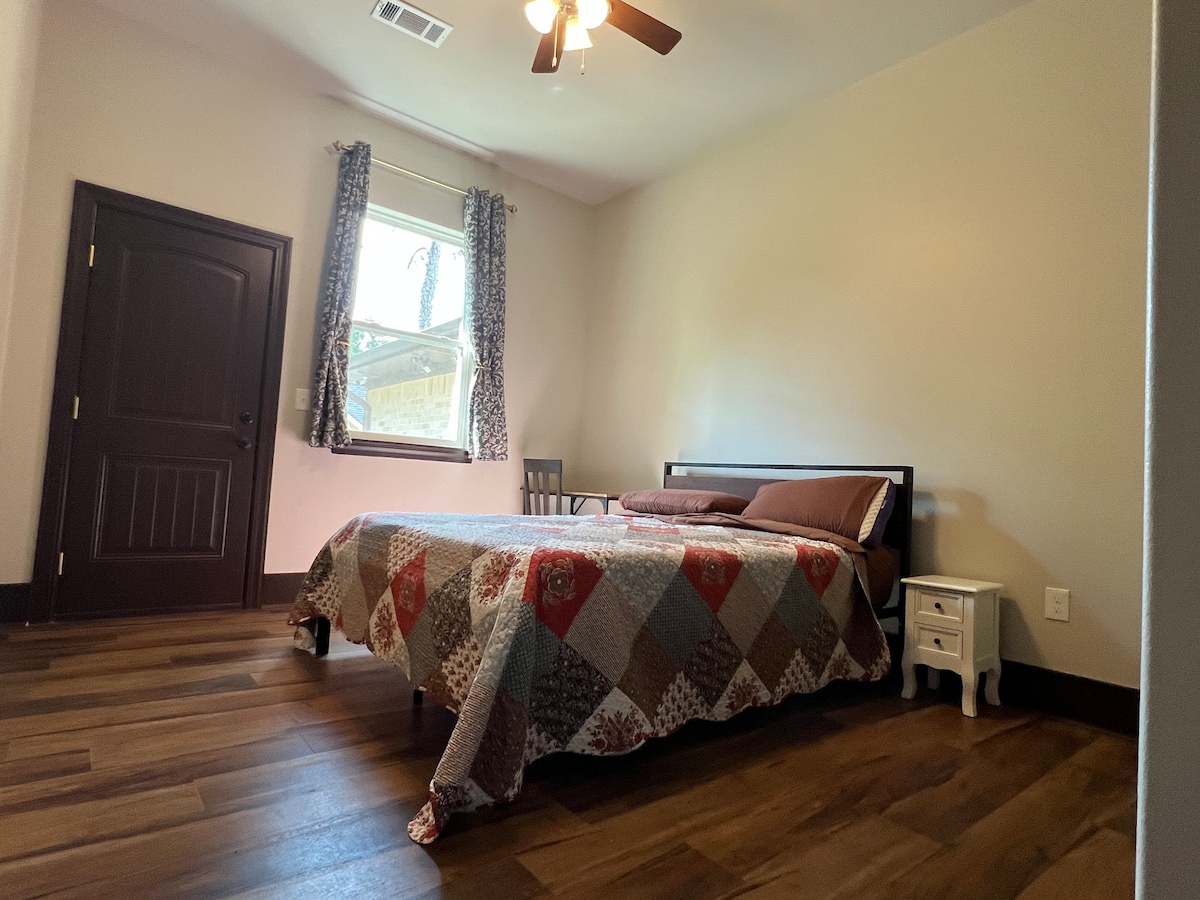 A cozy sleeping area features a queen bed dressed in a patterned quilt. A small bedside table stands beside the bed, and a wooden chair is positioned nearby. Natural light enters through a window adorned with patterned curtains, complementing the warm-toned wooden flooring.