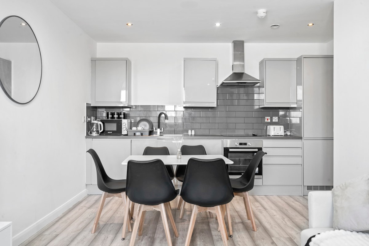 A modern kitchen area is displayed with sleek grey cabinets and stainless steel appliances. A dining table with black chairs is centrally positioned, accompanied by a large round mirror on the wall. Natural light brightens the space through the nearby window.