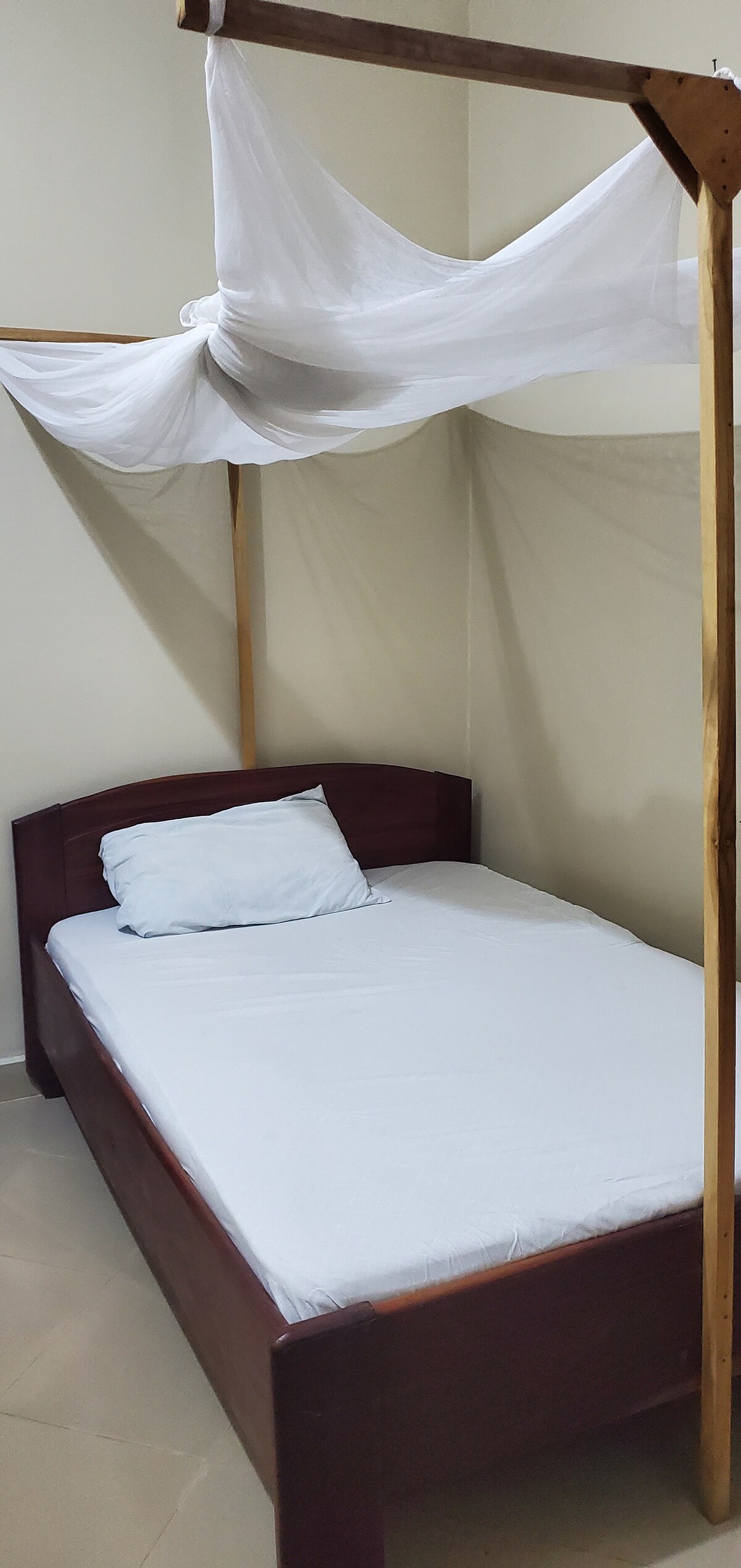 A single bed with a wooden frame is positioned in the room. A white mattress cover and a light blue pillow are present. A sheer white canopy drapes above the bed, adding a delicate touch to the space. Neutral walls provide a calm backdrop.