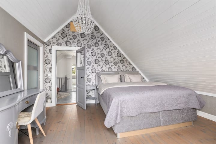 Une chambre avec du caractère!
Une chambre confortable avec un plafond en pente, du papier peint à fleurs et des planchers de bois clairs. Meublée avec un lit double, une coiffeuse et un lustre, une atmosphère confortable et élégante.