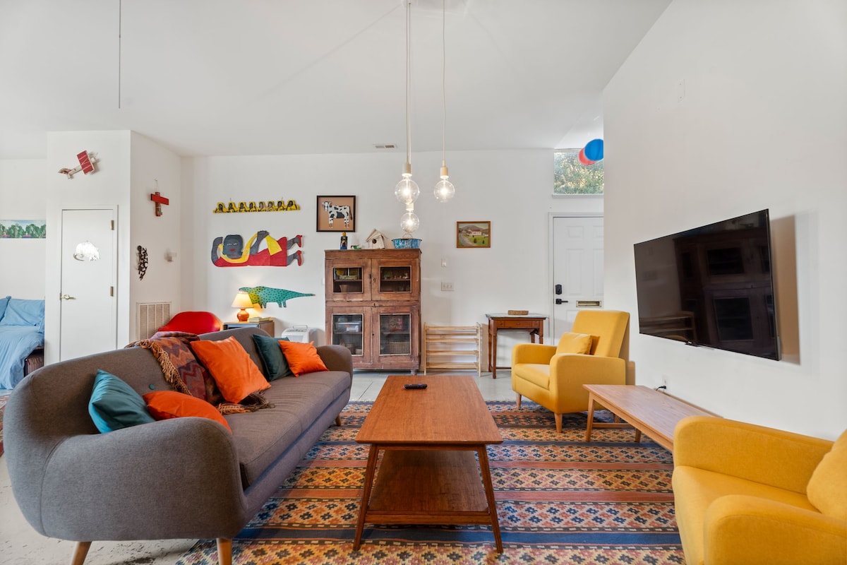 An open living area showcases a cozy couch with colorful cushions, flanked by two yellow chairs. A wooden coffee table is positioned in the center, surrounded by a patterned rug. A TV is mounted on the wall, and bright artwork adorns the space, enhancing the vibrant atmosphere.