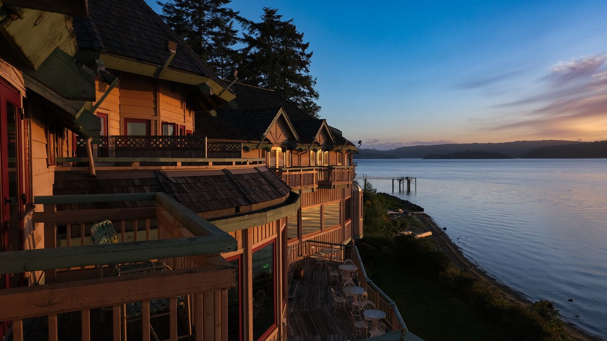 A scenic view of the lodge's exterior is captured at sunset, showcasing multiple balconies overlooking the water. Soft light reflects off the tranquil surface of the bay, while evergreen trees line the background, creating a serene setting.