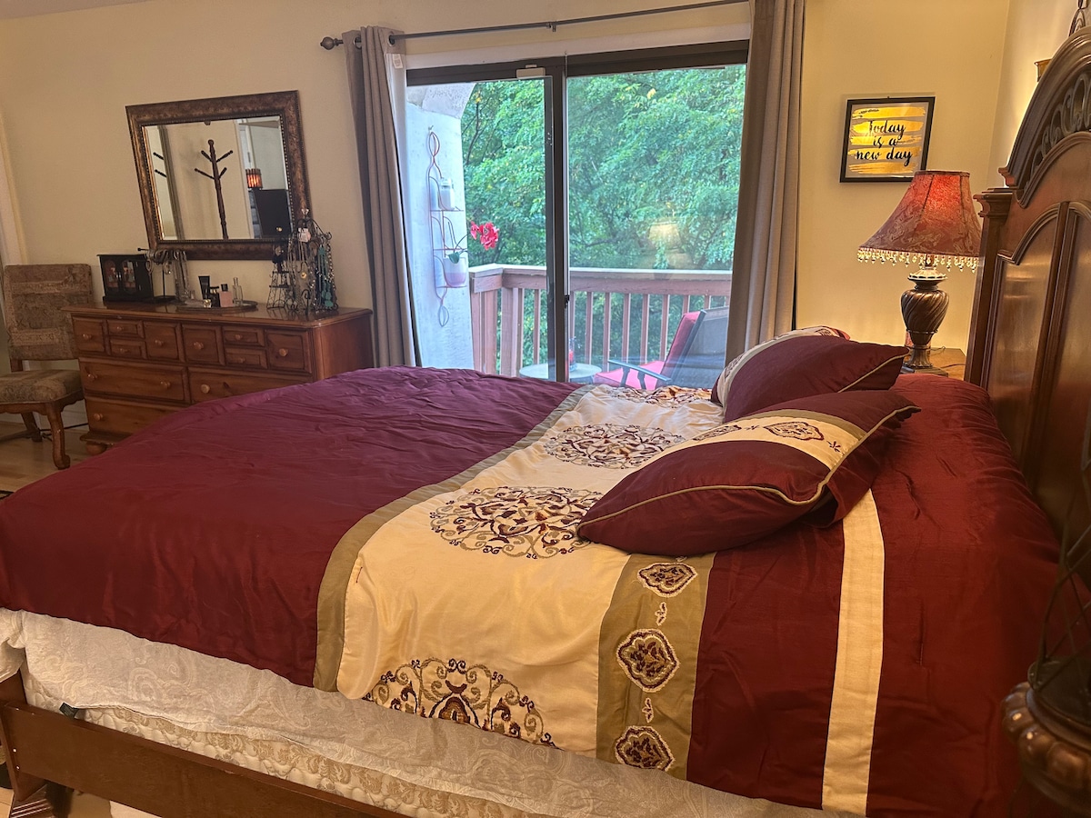 A spacious bed with rich burgundy and cream bedding is positioned in a well-lit master bedroom. A sliding glass door leads to a balcony with views of lush greenery. A wooden dresser and a mirror complement the room's decor, adding to its comfort.