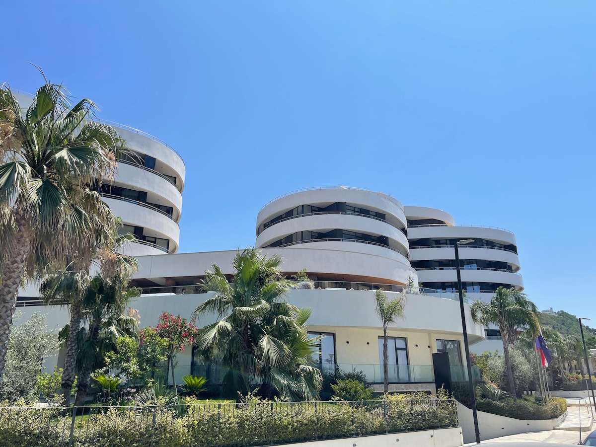 A modern architectural structure features curved balconies and large windows, surrounded by greenery including palm trees and shrubs. The building's clean lines and soft colors blend with the natural landscape, set against a clear blue sky.