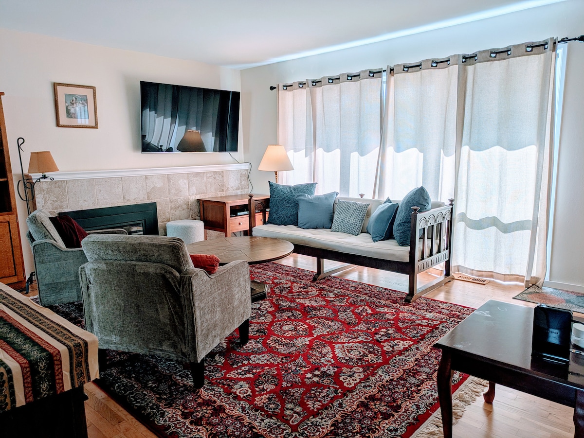 A comfortable living room features a large flat-screen TV mounted on the wall. A cozy seating arrangement includes a sofa and two armchairs, accompanied by a round coffee table. A patterned area rug adds warmth, while natural light filters through sheer curtains.