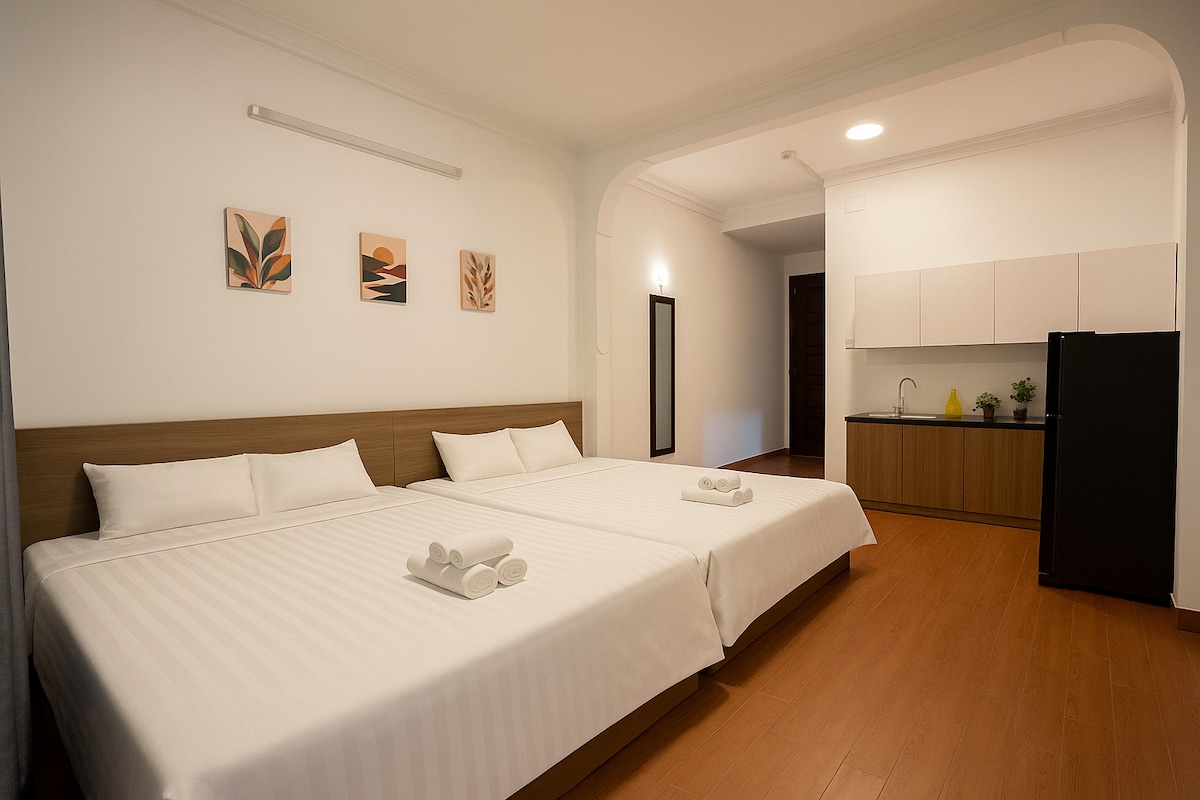 A spacious room features two queen-size beds with crisp white linens and neatly arranged towels. Above the beds, three pieces of wall art add subtle decoration. A kitchenette is visible in the background, equipped with a sink and a refrigerator, contributing to a functional living space.