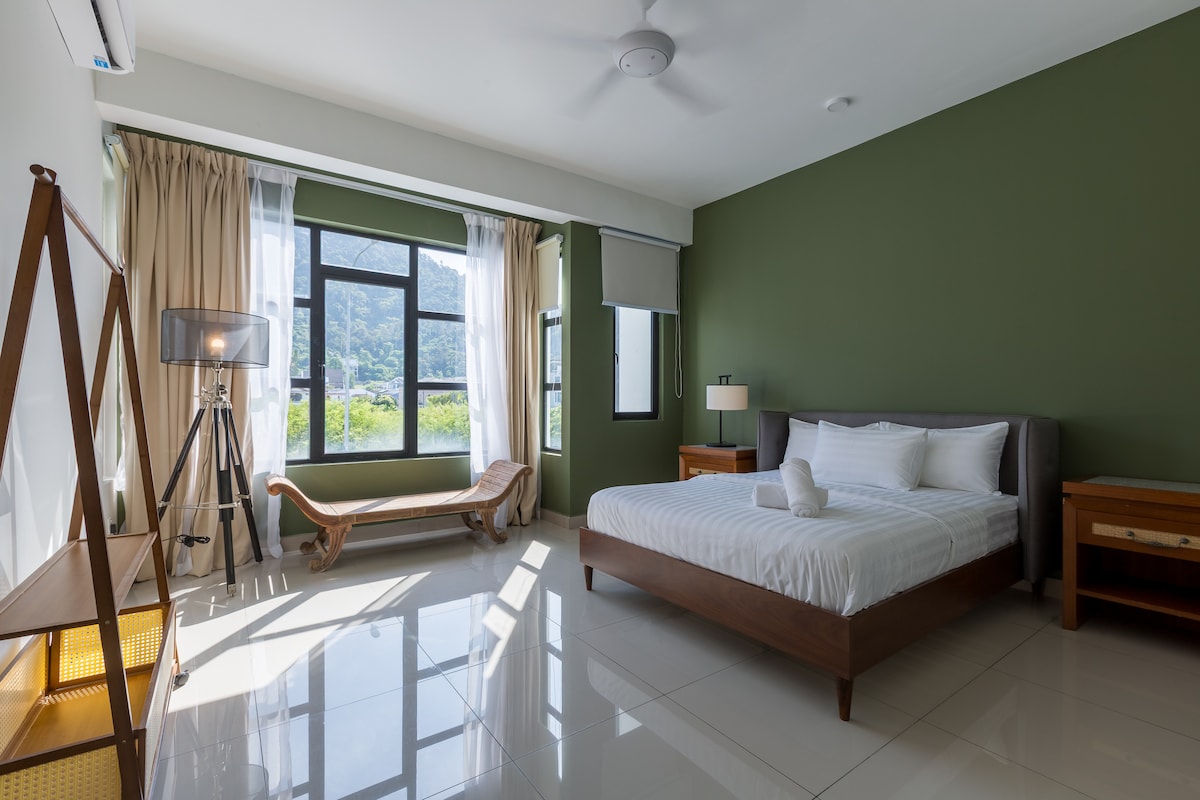 A bright bedroom features a spacious bed with crisp white linens, positioned under a ceiling fan. Natural light filters through large windows, framed by soft curtains. A vintage lounge chair and two wooden nightstands complement the contemporary décor, while green walls add a soothing touch.