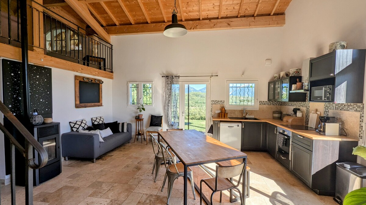 An open-concept living space features high wooden ceilings and large windows allowing natural light. A comfortable sofa is positioned near a dining table with metal chairs. A kitchen area is equipped with modern appliances, while a view of the surrounding landscape is visible through the sliding doors.