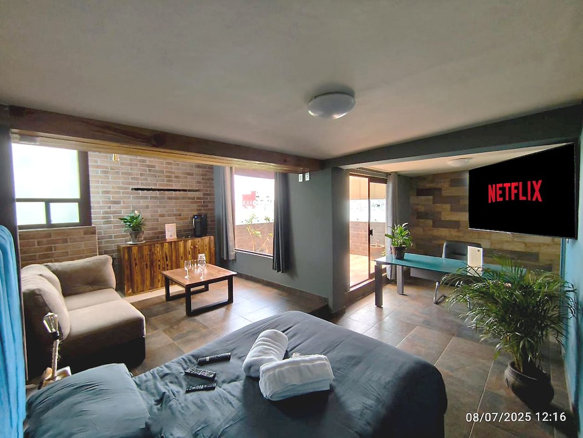 A spacious loft is equipped with a comfortable sofa and a coffee table, alongside a bed with fresh linens. Large windows allow natural light in, and a terrace can be seen through the glass doors. A wall-mounted screen is set up for streaming services.
