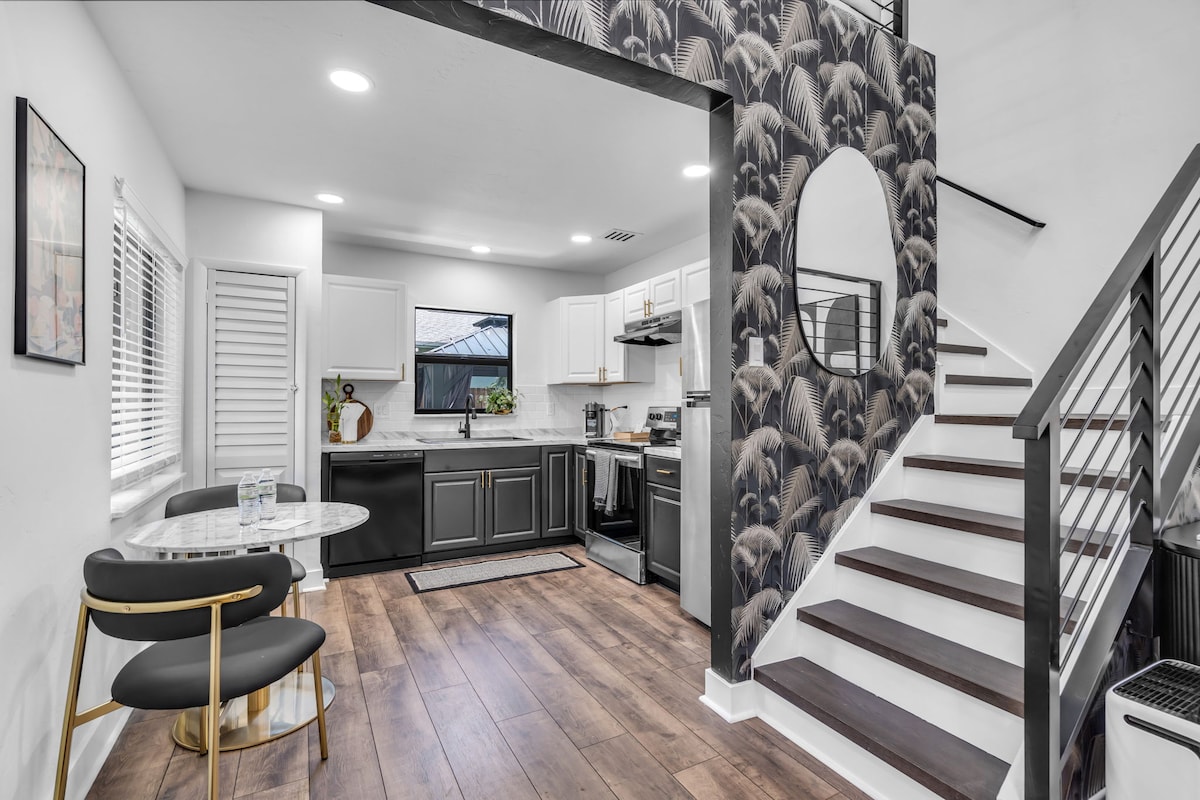 The kitchen and dining area display a modern design with sleek black cabinetry and a round dining table set for two. A spiral staircase is featured prominently, leading to the upper level, while a large round mirror reflects the stylish wallpaper adorned with palm leaves.