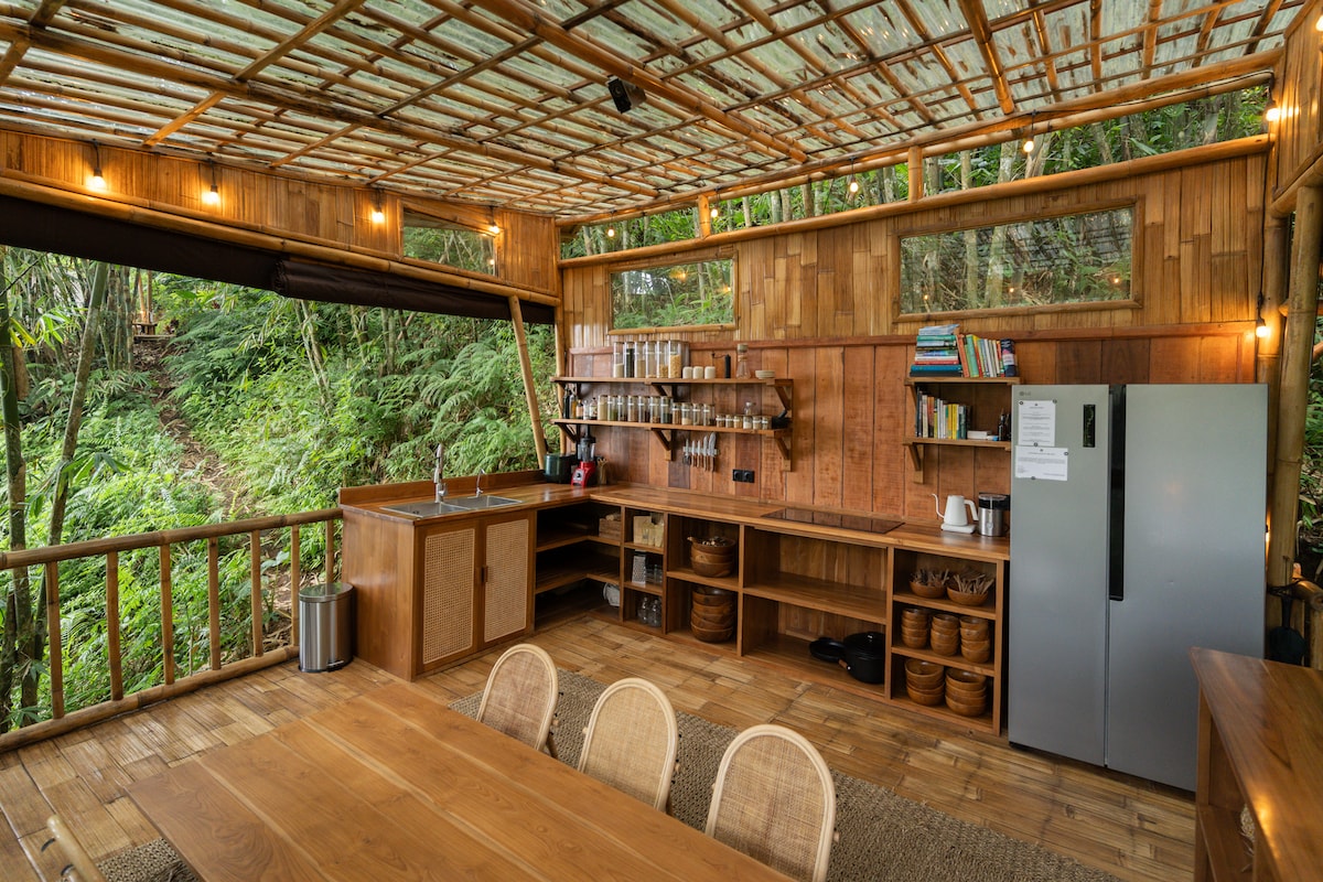 An open-air kitchen area is set against the lush greenery. Bamboo and wood materials create a warm and natural atmosphere. Features include a spacious dining table, a well-organized countertop with kitchenware, and a refrigerator. Large windows provide ample natural light and views of the surrounding forest.