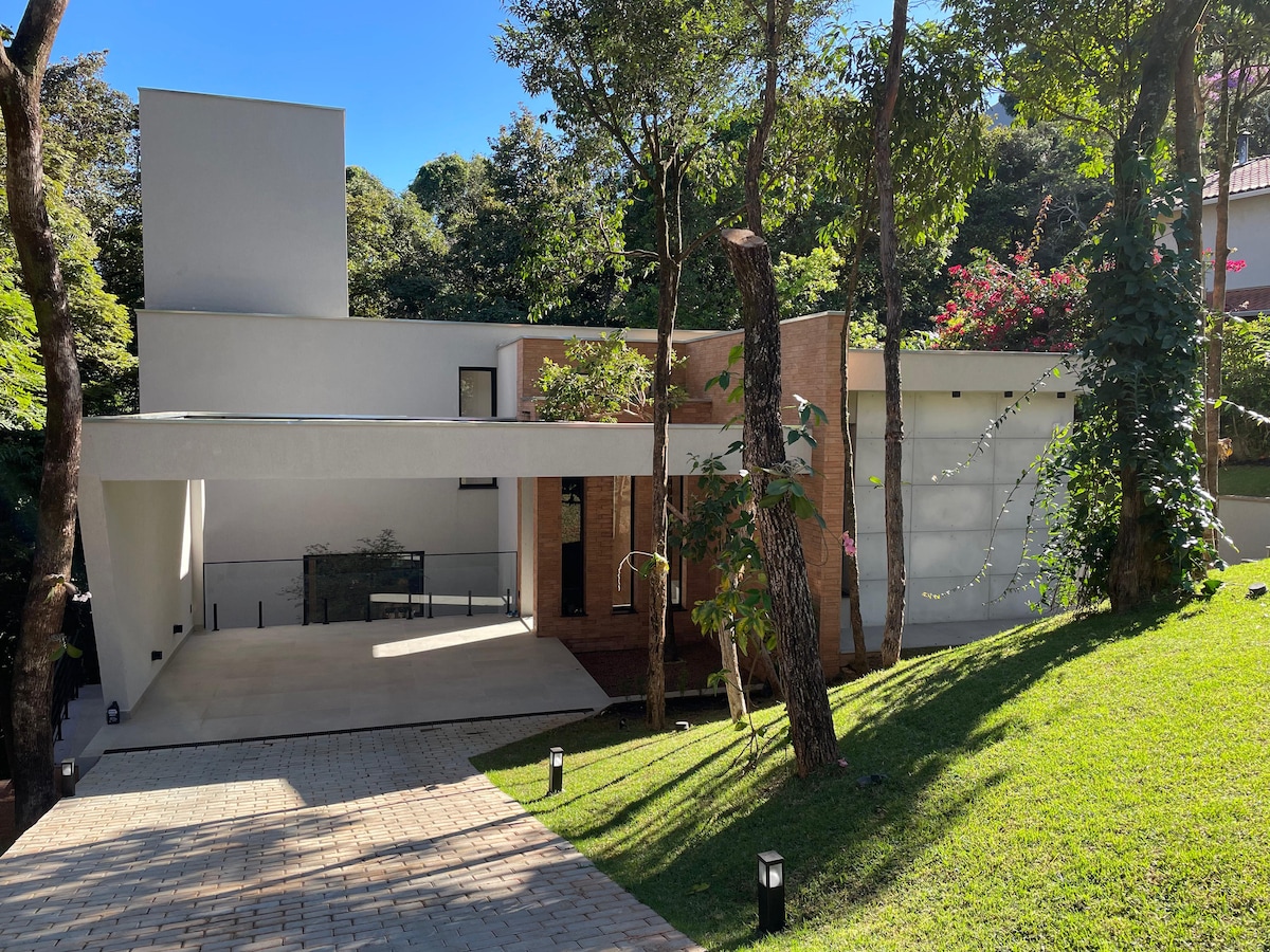 The exterior of a modern house is shown, surrounded by lush greenery. A paved driveway leads to the entrance, flanked by mature trees. The structure combines smooth white walls with natural wood accents, creating a harmonious integration with the landscape.