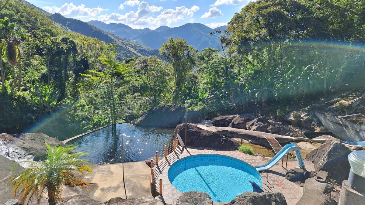 A landscape showcases a natural pool surrounded by lush greenery and rocky formations. A smaller circular pool is visible, featuring a water slide. Majestic mountains form the backdrop under a clear blue sky, enhancing the scenic atmosphere.