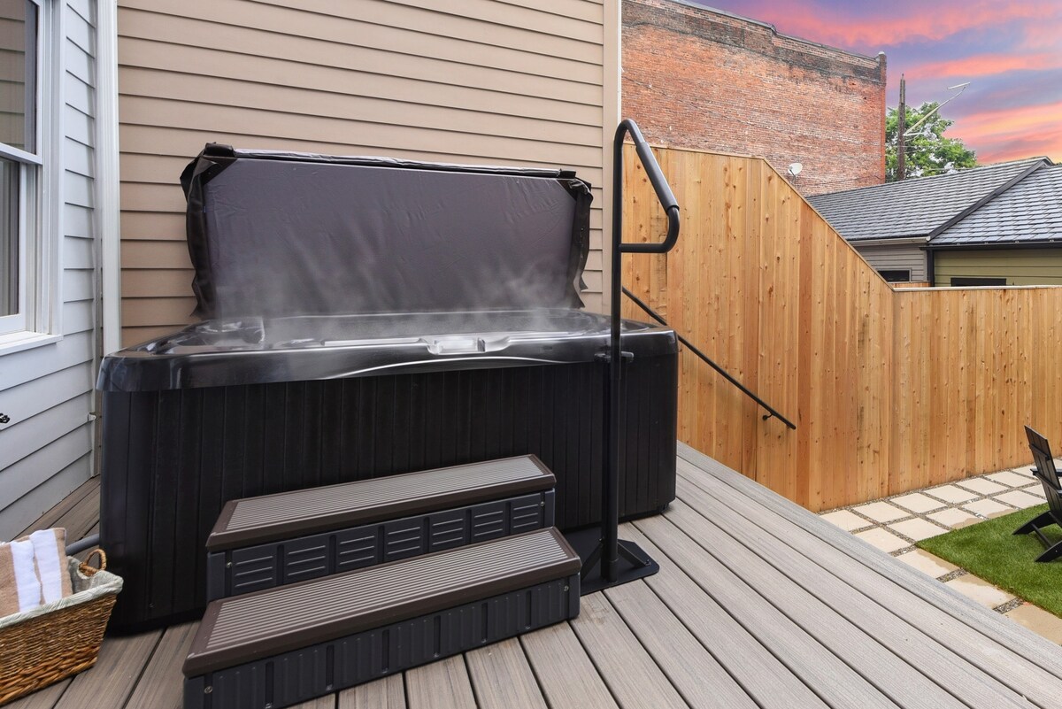 A spacious hot tub is positioned on a wooden deck, surrounded by a privacy fence. Steps lead up to the tub, where steam rises gently, hinting at inviting warmth. The backdrop features a setting sun and a hint of brick wall, creating a serene atmosphere.