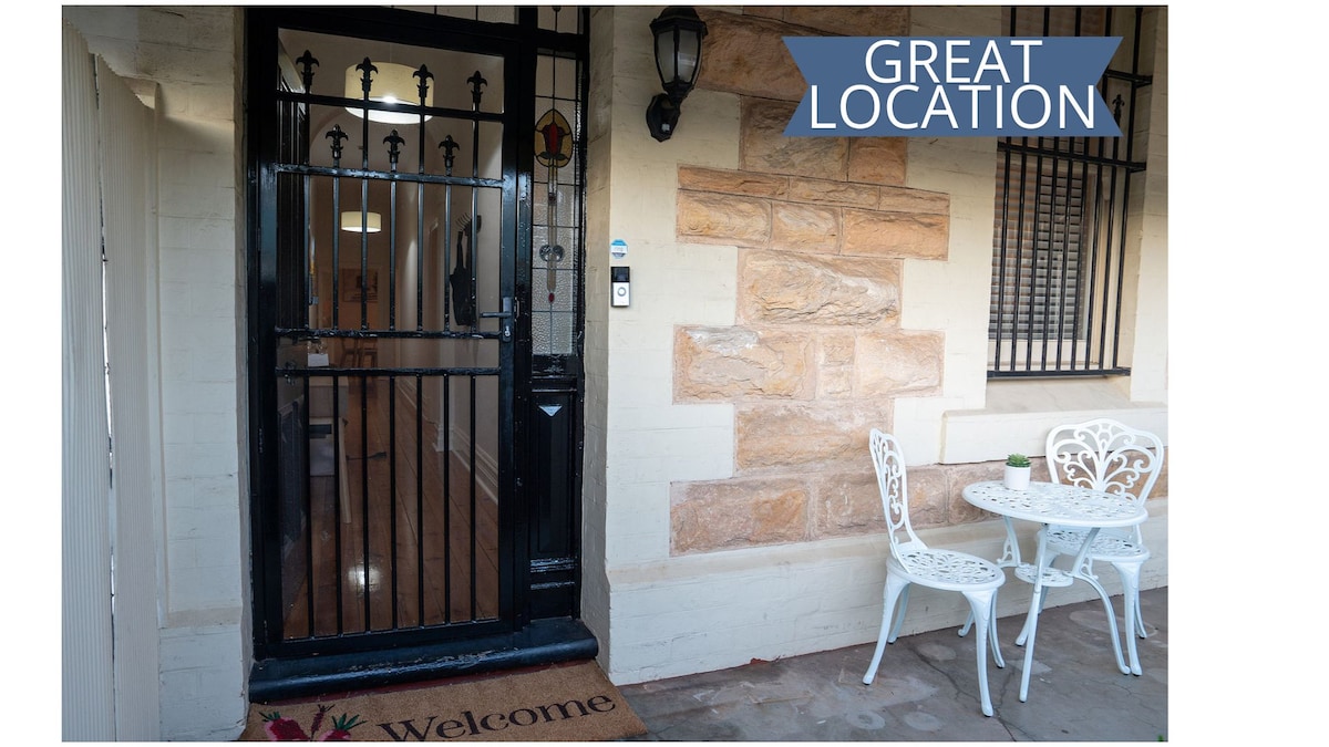 The entrance of The Arches features a decorative black iron gate and a warm welcome mat. A small outdoor seating arrangement includes two white chairs and a round table, positioned against a backdrop of textured stone walls. Bright light filters through the nearby windows.