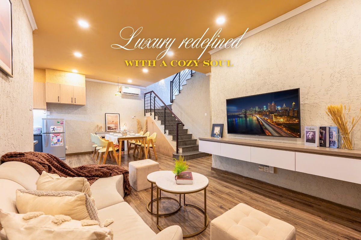 A spacious living area is featured, showcasing a cozy sofa with a warm blanket draped over it. A modern dining space with a wooden table is visible in the background. A large TV mounted on the wall displays a scenic city view, adding to the inviting atmosphere.