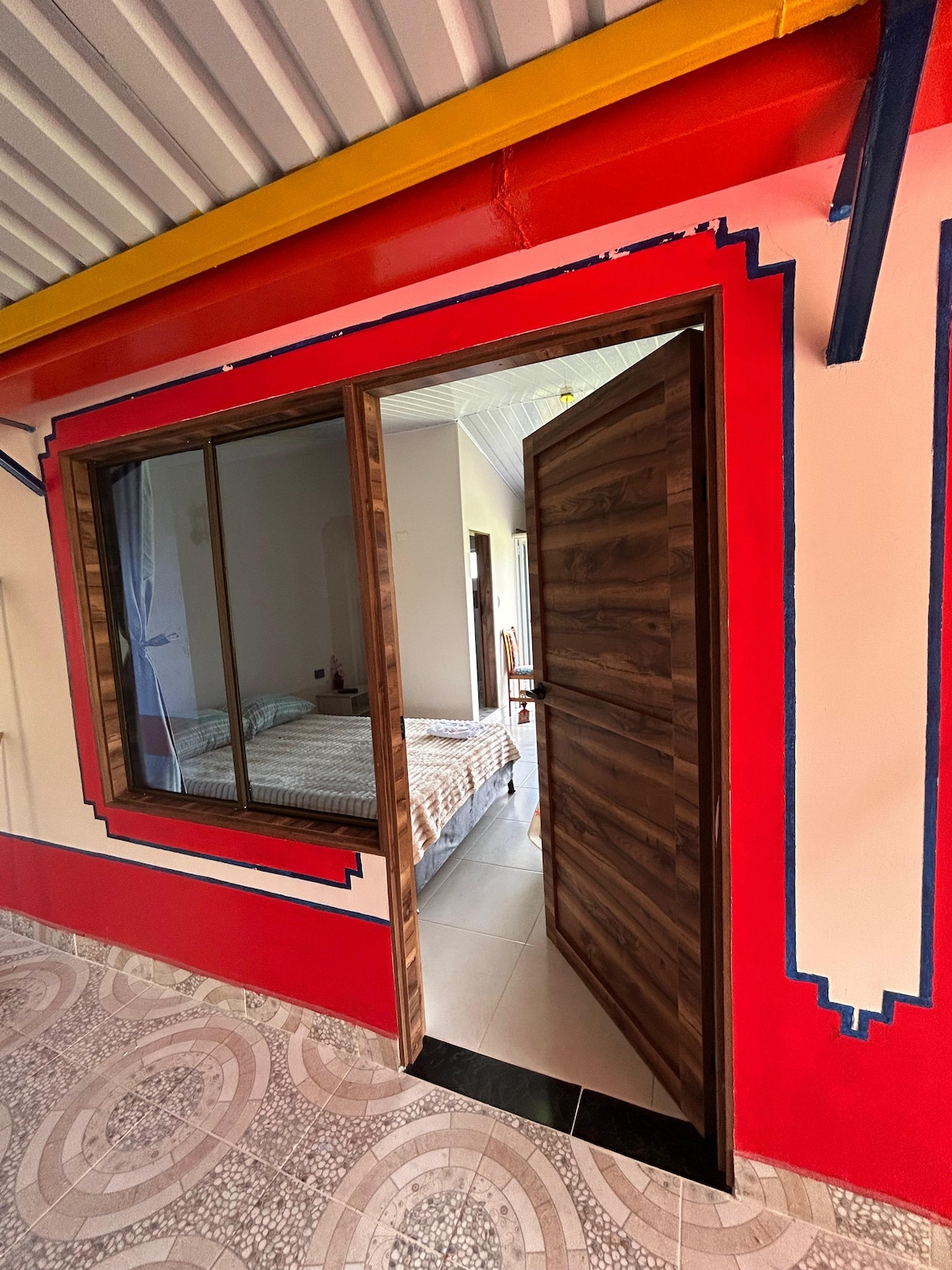 A doorway opens into a warmly decorated bedroom, featuring a spacious bed with light bedding. Colorful murals accent the surrounding walls, and natural light floods through large windows. The floor is composed of circular patterned tiles, creating a harmonious blend with the room's vibrant design.