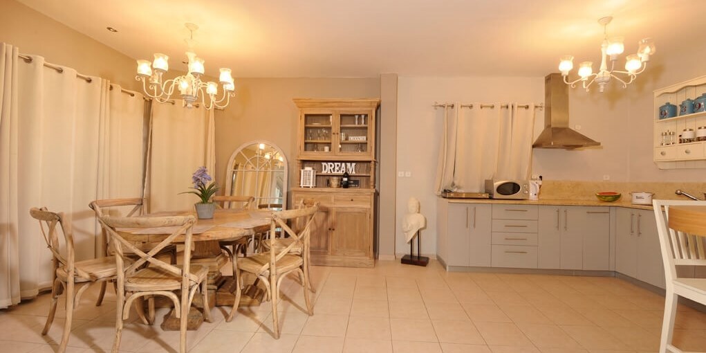 A well-lit dining area features a round wooden table surrounded by six chairs. A stylish cabinet holds various kitchen items, while a countertop with appliances is visible. Soft lighting from chandeliers enhances the ambiance, and window drapes offer privacy.