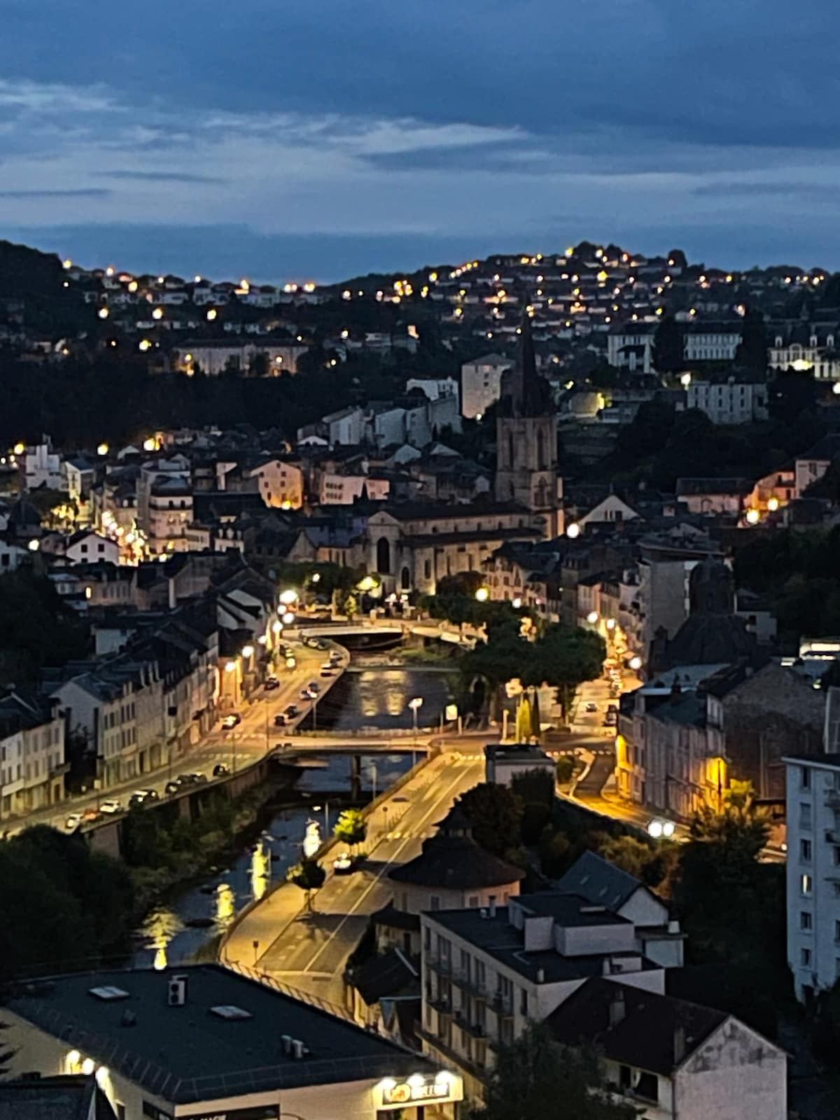 An evening view captures the cityscape of Tulle, illuminated by warm streetlights. The winding river reflects the glow, while a church steeple rises prominently among the buildings. The surrounding hills are softly lit, offering a serene ambiance to the scene.