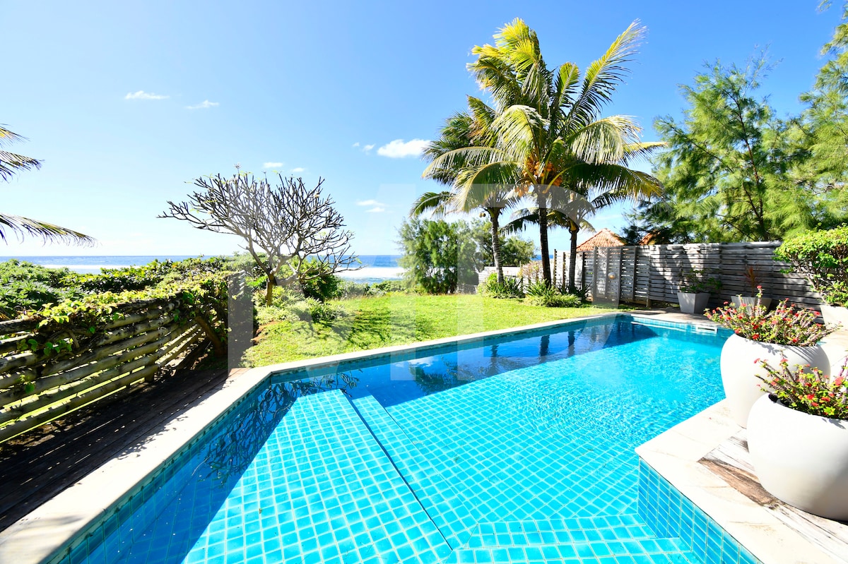 A private pool is surrounded by lush greenery and palm trees, highlighting a tranquil outdoor space. The clear blue water of the pool reflects the bright sky, and the ocean is visible in the background, enhancing the serene atmosphere of the setting.