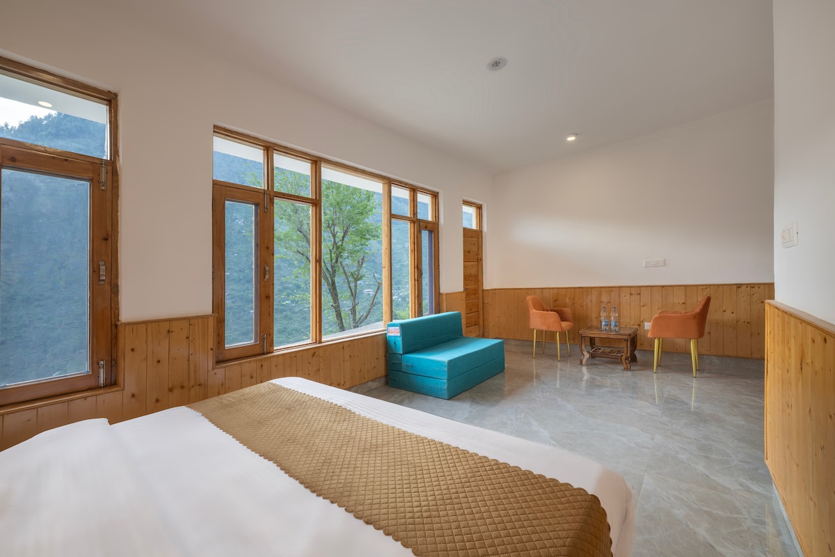 A spacious room features large windows offering views of the mountain valley. Wooden paneling lines the walls, and a comfortable bed is complemented by a teal sofa and two orange chairs. A small table is situated between the seating area, creating a cozy retreat.