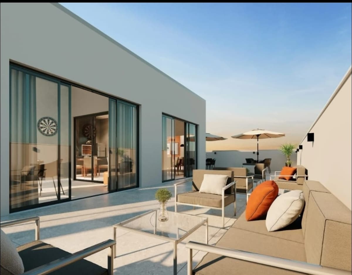 An outdoor terrace is featured, furnished with a glass table and comfortable seating arrangements. Sun loungers and a shaded umbrella are visible. Large glass doors lead to the interior, allowing for a seamless transition between indoor and outdoor spaces.