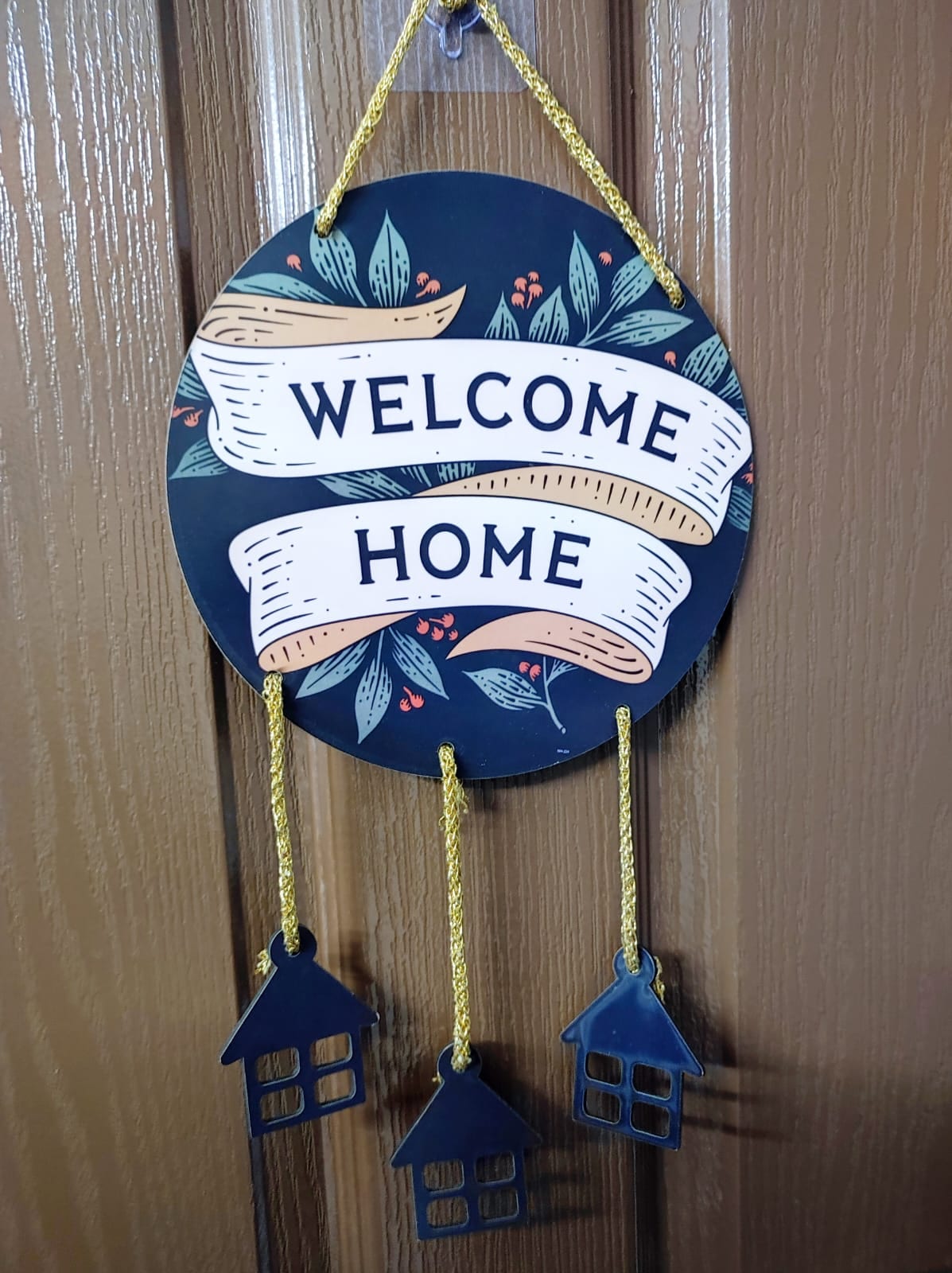 A circular welcome sign hangs prominently on a wooden door, featuring the words 'WELCOME HOME' in bold lettering. Decorative elements include floral designs, with small house-shaped charms dangling beneath the sign, adding a charming touch to the entryway.