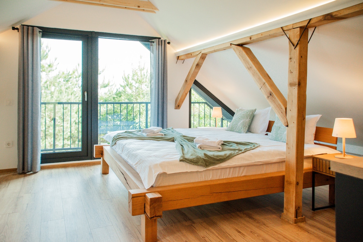 A spacious bedroom features a large wooden bed with white linens and soft green accents. Natural light enters through a large window with balcony access, framed by light curtains. The room includes wooden flooring and exposed wooden beams, contributing to a modern, yet cozy environment.