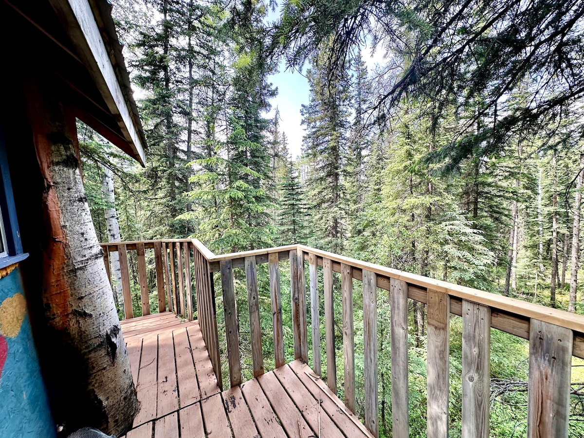 A wooden deck wraps around the treehouse, offering a serene view of surrounding pine trees and lush greenery. The deck features slatted wood flooring, providing a rustic touch and an inviting space for relaxation.