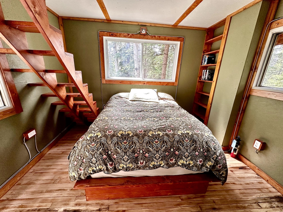 An interior view of the treehouse featuring a cozy double bed adorned with a patterned blanket. Surrounding windows allow for natural light, revealing wooden floors and walls in calming green tones. A staircase leads to an upper level, while a small bookshelf is positioned nearby.