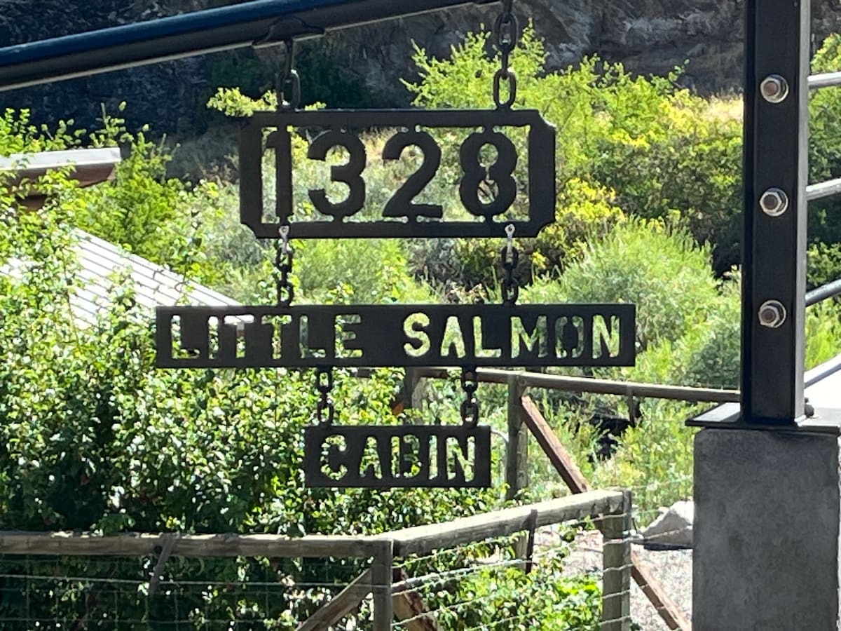 A rustic metal sign displays the address '1328' and the name 'Little Salmon Cabin.' The sign is suspended from a structure, surrounded by lush greenery and natural elements, providing a welcoming entrance to the cabin.
