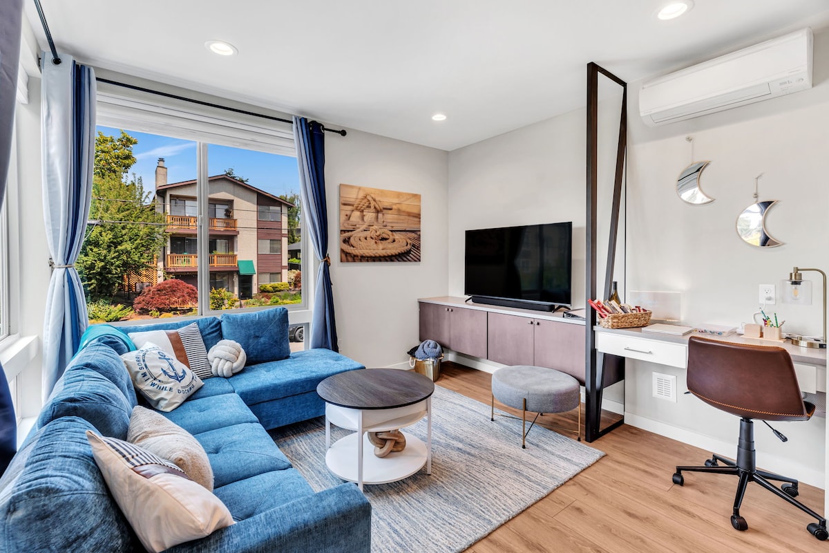 A modern living room features a plush blue sectional sofa and a round coffee table atop a soft area rug. Natural light filters through large windows, highlighting a wall-mounted television and a sleek desk with a basket. Decorative accents and greenery enhance the space.