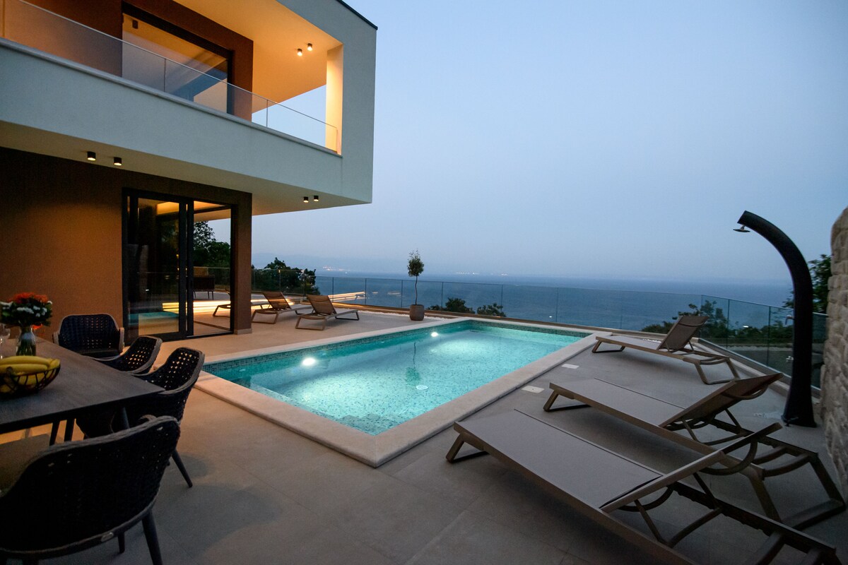 An outdoor space features a private pool surrounded by sun loungers. A dining table is visible, set for outdoor meals. Large glass doors open to the villa, providing views of the sunset over the sea.