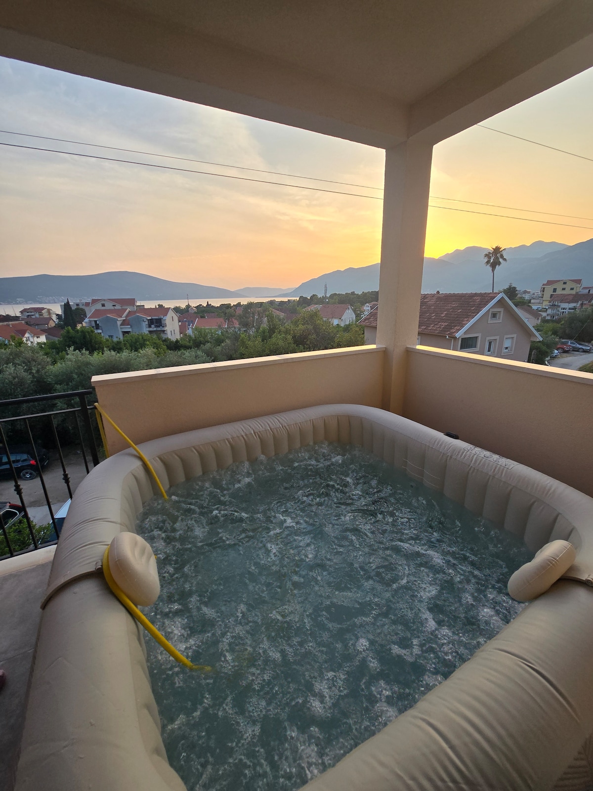 An inflatable Jacuzzi is situated on the terrace, providing a serene setting for relaxation. The view includes distant mountains and a soft sunset glow, creating a peaceful atmosphere. The terrace is enclosed with a railing, ensuring safety while enjoying the outdoor space.