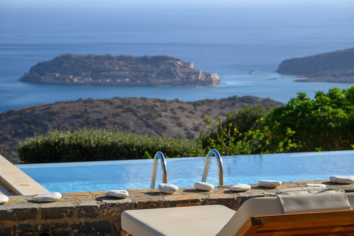 An infinity pool stretches toward the horizon, framed by a stone deck adorned with smooth pebbles. Vibrant waters merge with the expansive sea view, revealing an island in the distance. Lush greenery surrounds the area, emphasizing the serene landscape.