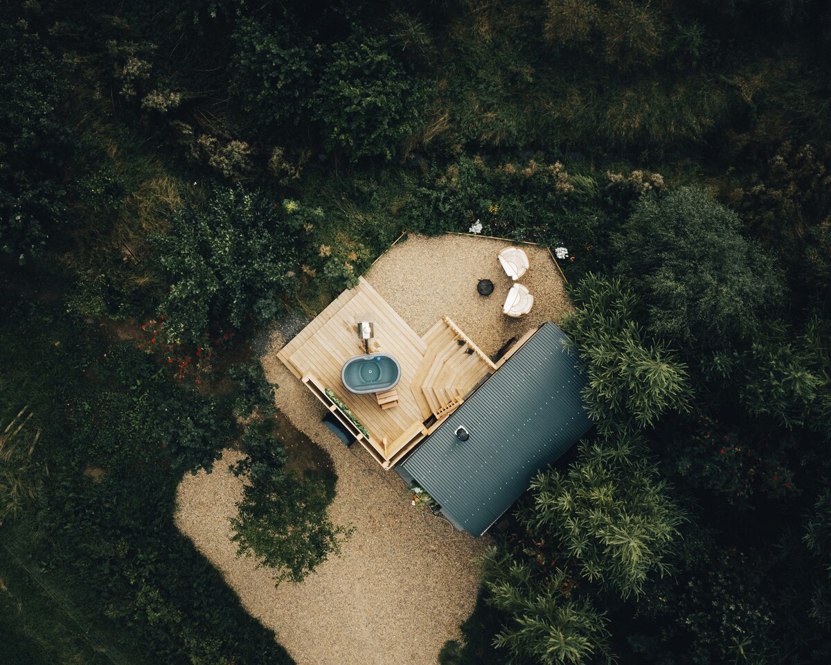 An aerial view showcases the shepherd's hut nestled amidst greenery. A wooden deck features a hot tub and lounge area, surrounded by natural landscaping. Gravel paths weave through the outdoor space, leading to comfortable seating arrangements for relaxation.