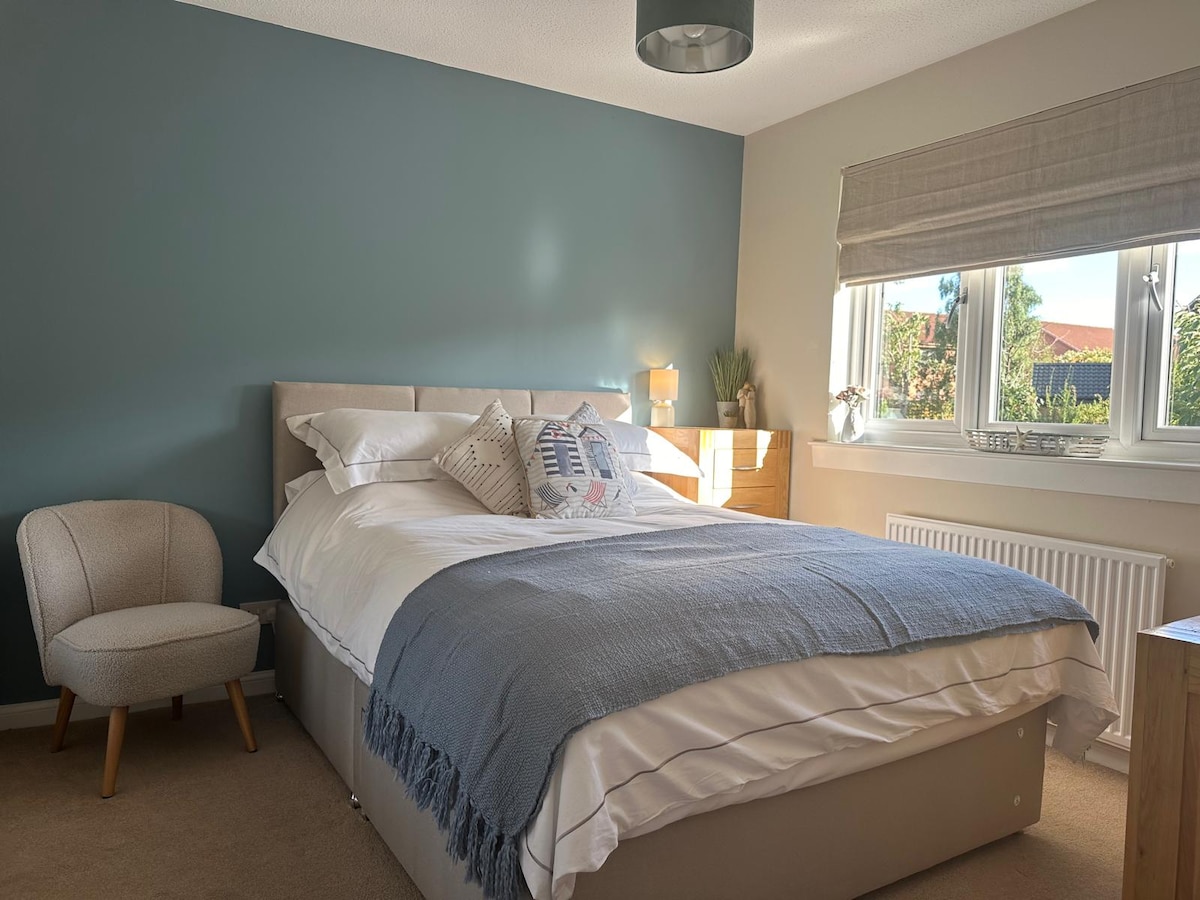 A modern double bedroom is presented with a spacious bed, adorned with a light blanket and decorative pillows. Natural light pours in from the window, casting a soft glow across the room. A comfortable chair and bedside tables complement the decor.