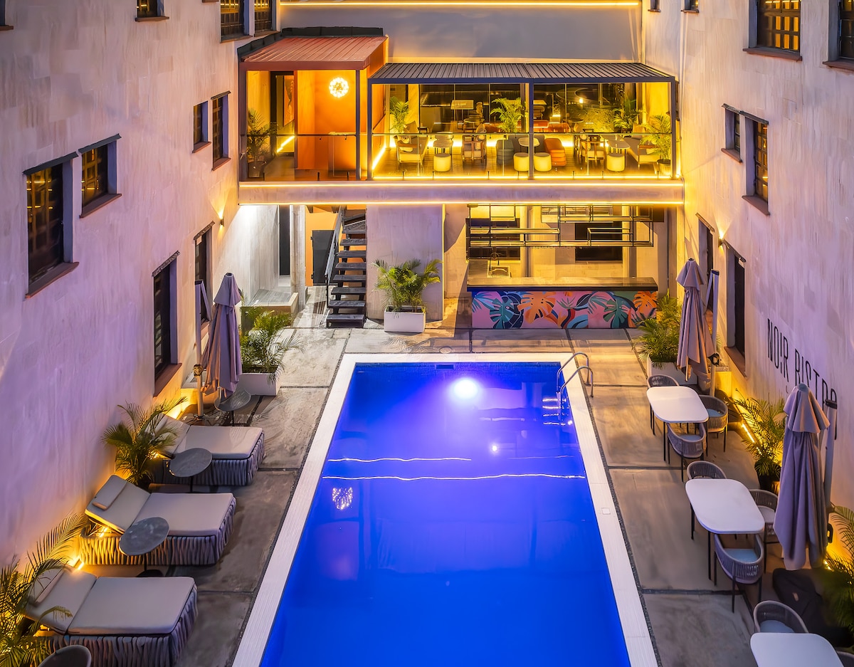 A spacious outdoor area is highlighted by a vibrant blue pool, surrounded by lounge chairs and dining tables. Soft lighting illuminates the poolside, while a balcony above showcases an inviting seating area. Lush greenery contributes to a relaxing ambiance.